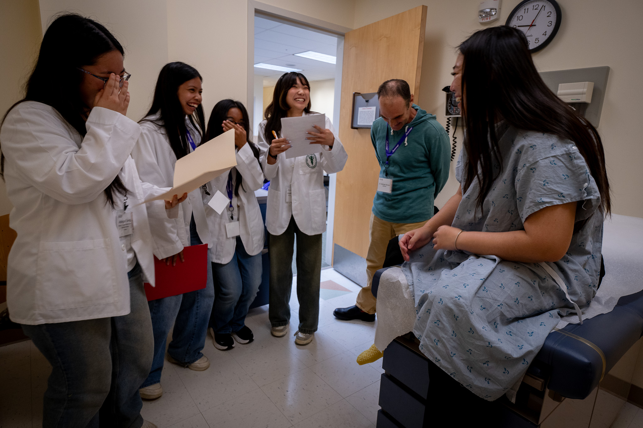 La estudiante de medicina Darcy Tokunaga, a la derecha, se ríe con los estudiantes de Kaiser High School mientras interpreta a una paciente durante una sesión de aprendizaje en JABSOM el 7 de abril de 2026. De izquierda a derecha están Abbigail Song, Kailie Arpin, Mia Meeds, Kaitlin Miyashiro y el Dr. Michael Wyman. El evento y la competencia incluyeron mesas de autopsias virtuales creadas por Anatomage de Santa Clara, California (Craig Fujii/Civil Beat/2026).