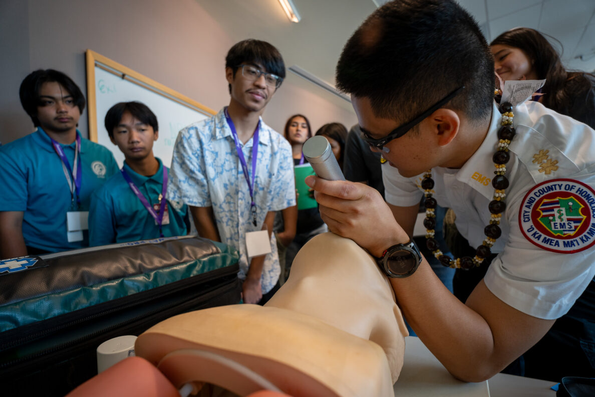 El EMT Cameron Hara muestra a los estudiantes cómo colocar un tubo de respiración en un maniquí de vías respiratorias durante una sesión educativa en JABSOM el 7 de abril de 2026. Desde la izquierda, Jayden Cabel-Suehiro, Jaiden Aquino y Zander Munalem observan.
