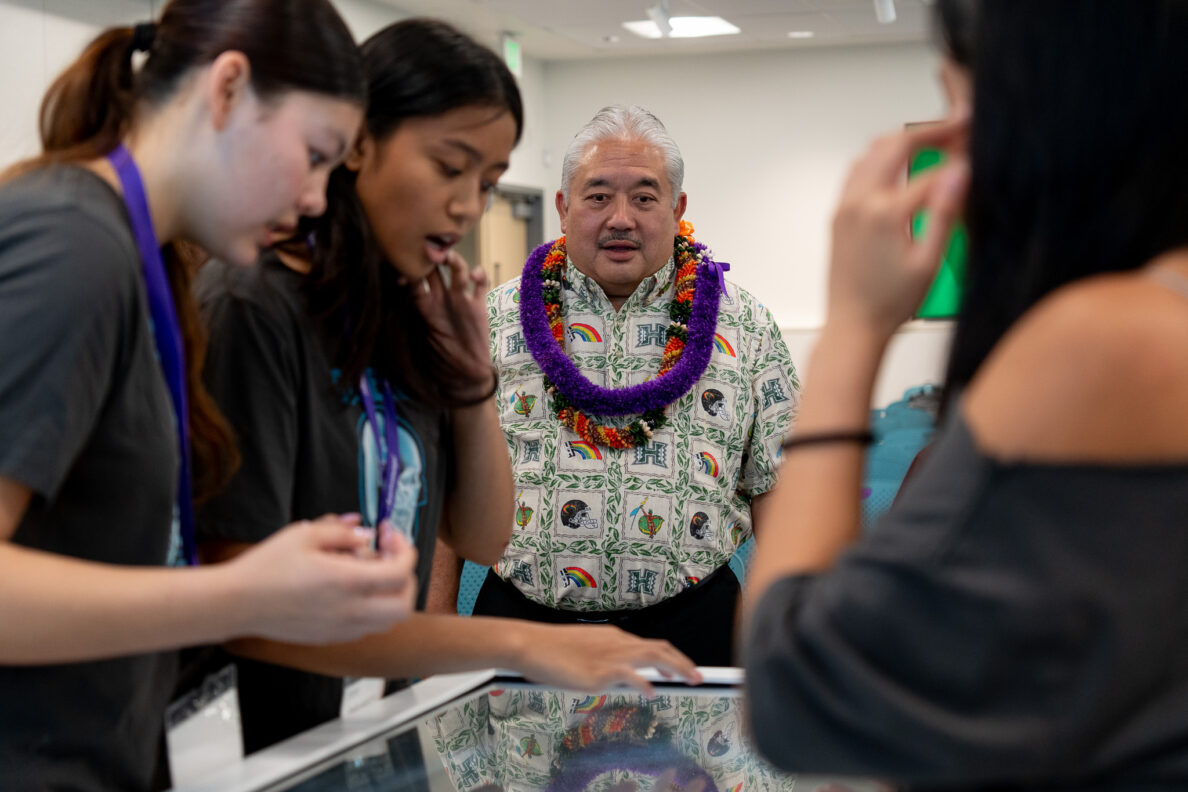 Un hombre con una camisa hawaiana y lei observa a un estudiante con una camiseta gris trabajar en una máquina de mesa.