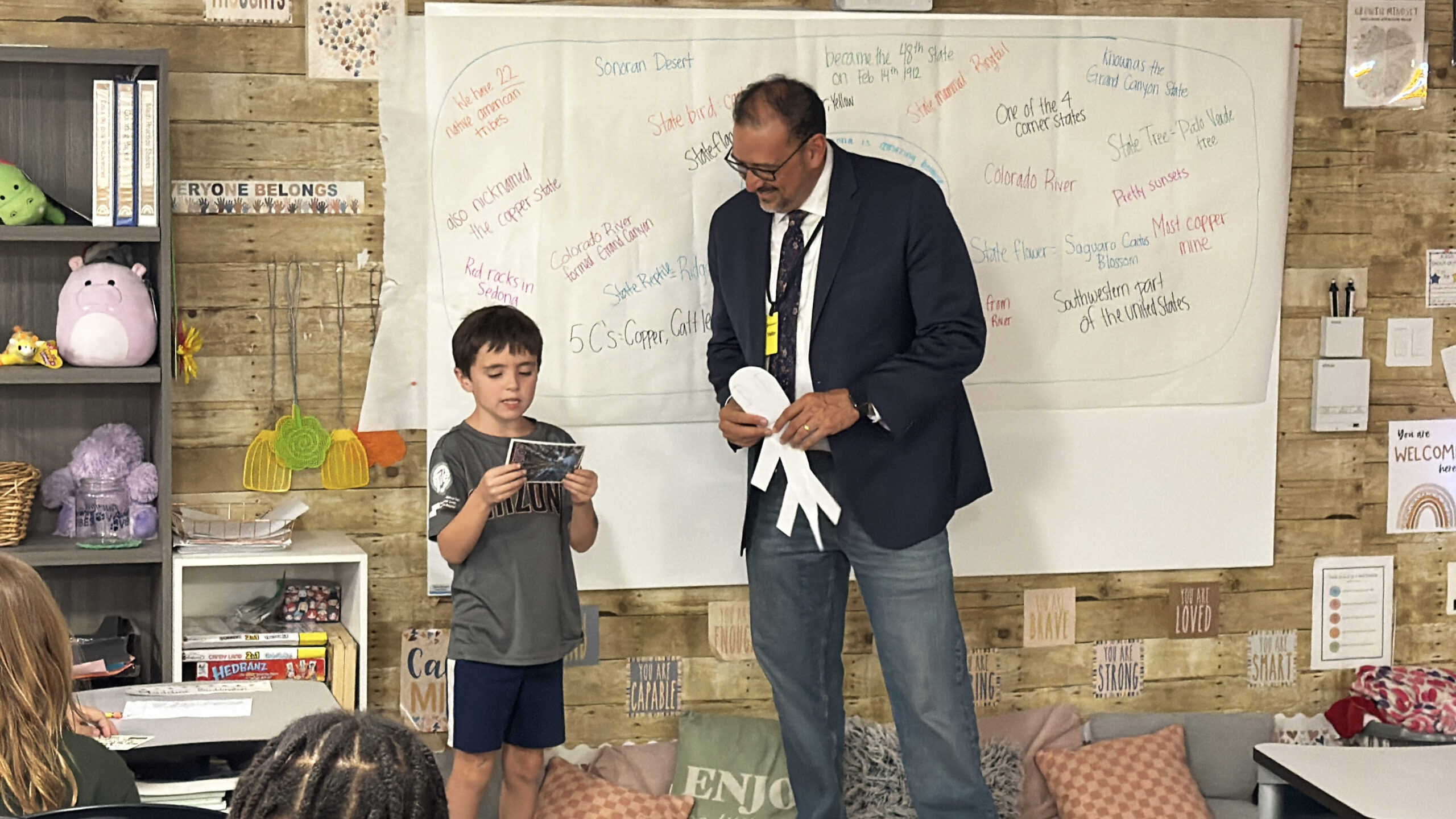 Un niño al frente del salón de clases comparte una postal con el presidente del comité Arizona America250, Adrián Fontes.